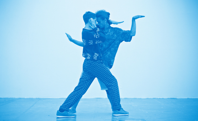 Två personer dansar uttrycksfullt nära varandra med ingenting utom en ljus vägg i bakgrunden. Hela bilden är tintad med ett blått skimmer.