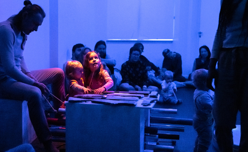 Två små barn håller varsin hand på ett bord eller något som skulle kunna vara ett slagsverksinstrument. Runt omkring dem sitter ett fler vuxna och barn, varav en av de vuxna också spelar ett slagsverksinstrument. En spotlight riktar uppmärksamheten mot de två barnen.
